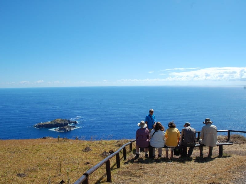 Tourists sitting on a wooden bench with their guide explaining. The place overlooks an islet and the blue Pacific Ocean at Orongo archaeological site (Easter Island)