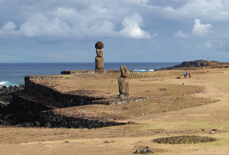 Two moais statues standing in Tahai archaeological site on Easter Island having the blue Pacific Ocean as backdrop (and travelers contemplating the one with a hat)