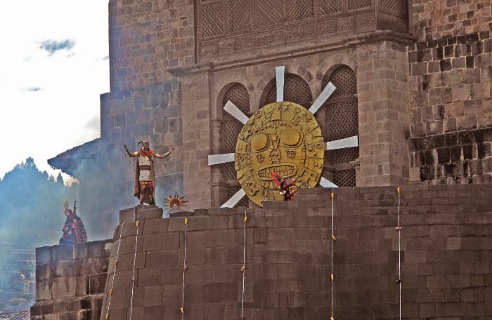 A man standing and opening arms looking at the sky with a stone building and a symbol of a sun as backdrop