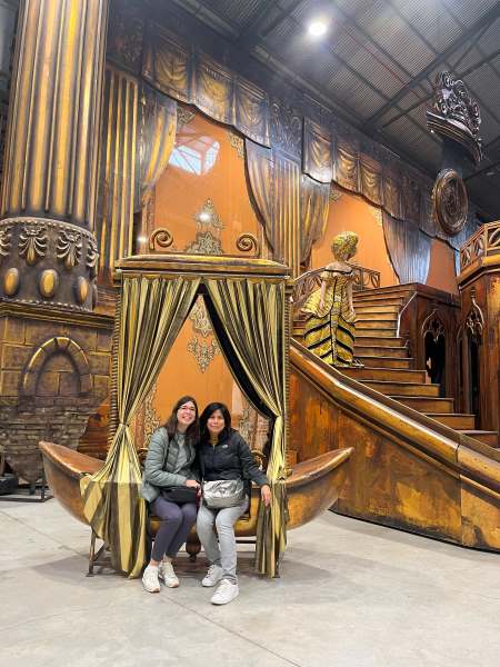 Two women sitting in a sort of tub with golden curtains as frame and stairs and a column as backdrop